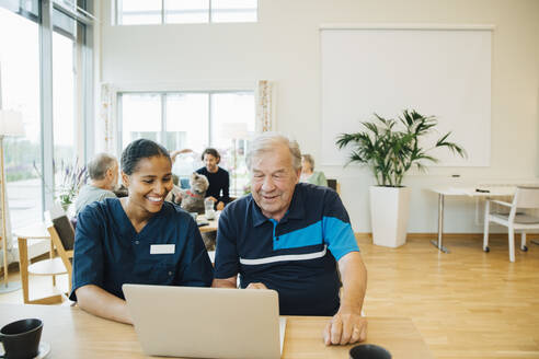 Lächelnde junge Krankenschwester, die einem älteren Mann bei der Benutzung eines Laptops am Esstisch hilft - MASF16235