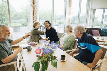 Lächelnder Mann inmitten älterer Menschen, die am Esstisch vor dem Fenster eines Pflegeheims sitzen - MASF16241