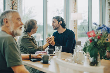 Senior woman talking with grandson while sitting by man at dining table in nursing home - MASF16242