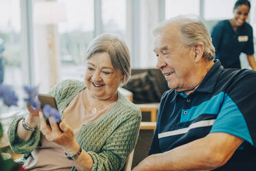 Smiling senior woman sharing smart phone with man while sitting at retirement home - MASF16243