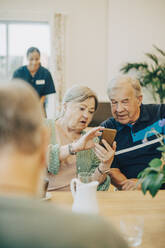 Ältere Frau teilt ihr Smartphone mit einem Mann, während sie am Esstisch im Altersheim sitzt - MASF16244