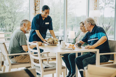 Lächelnde junge Mitarbeiterin des Gesundheitswesens serviert älteren Männern und Frauen, die am Esstisch sitzen, das Frühstück - MASF16245