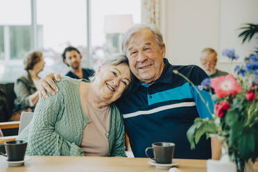 Porträt älterer Freunde, die am Esstisch eines Pflegeheims sitzen und sich umarmen - MASF16248
