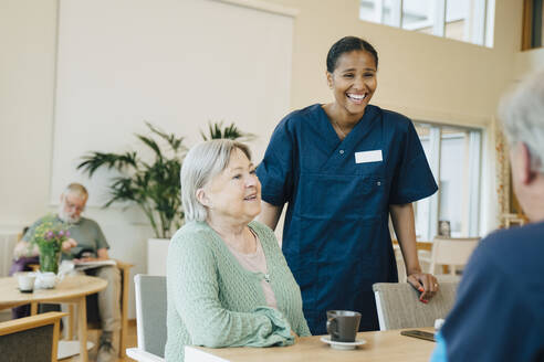 Fröhliche junge Krankenschwester, die bei einer älteren Frau steht und einen Mann im Altersheim betrachtet - MASF16253