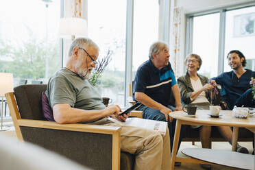 Älterer Mann, der sein Smartphone benutzt, während er mit einer Zeitung auf einem Sessel sitzt, der von Freunden und dem Pfleger in einem Altenpflegeheim aufgestellt ist - MASF16254