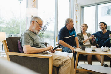 Senior man using smart phone while sitting with newspaper on armchair by friends and caretaker at elderly nursing home - MASF16254