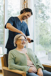 Lächelnder Pfleger beim Bürsten der Haare einer älteren Frau im Altersheim - MASF16264