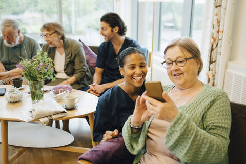 Ältere Frau im Ruhestand teilt ihr Smartphone mit einer lächelnden jungen Krankenschwester bei einem Gemeindeeinsatz, lizenzfreies Stockfoto