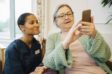 Lächelnde Pflegerin betrachtet ältere Frau, die im Pflegeheim ein Mobiltelefon benutzt - MASF16268