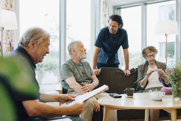 Male healthcare worker talking to senior man sitting on sofa amidst friends at retirement home - MASF16272