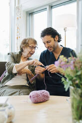 Senior woman teaching knitting to male nurse while sitting on sofa at elderly care home - MASF16273