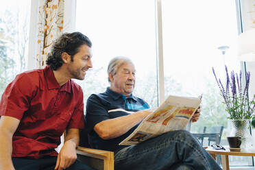 Lächelnde Pflegerin sitzt neben einem älteren Mann, der am Fenster eines Altenheims Zeitung liest - MASF16278