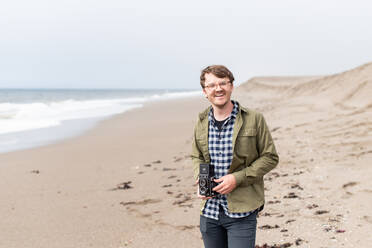 Portrait of young man laughing while holding film camera on beach - CAVF72593