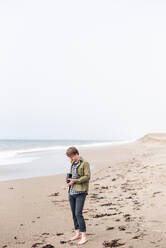 Young man standing on beach taking photograph with film camera - CAVF72594