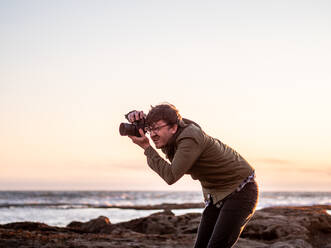 Mann hockt und fotografiert mit Digitalkamera an der Küste - CAVF72602