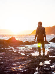 Man standing on rocky shore facing ocean with sun setting - CAVF72603