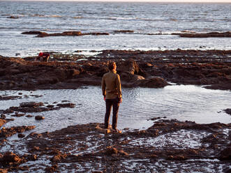 Man standing at edge of tide pool looking out toward sunset - CAVF72606