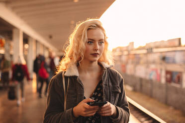 Junge Frau mit Smartphone in der Hand beim Warten auf den Zug - CAVF72627