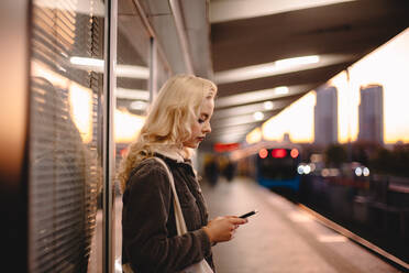 Junge Geschäftsfrau, die ihr Smartphone benutzt, während sie auf einen U-Bahn-Zug wartet - CAVF72643