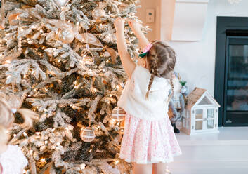 Kleinkind in rosa Kleid setzen ein Ornament auf einem beflockten Weihnachtsbaum - CAVF72649