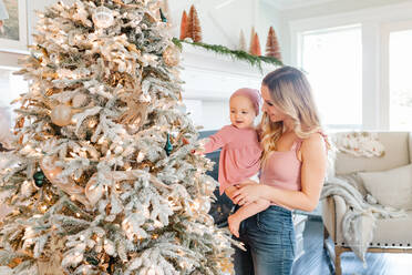 Mother Holding baby daughter by christmas tree - CAVF72657