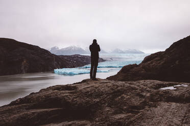 Mann stehend und fotografierend auf einem Felsen am See mit Gletschern - CAVF72698
