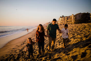 Vierköpfige Familie hält sich an den Händen, Spaziergang am Strand bei Sonnenuntergang - CAVF72750