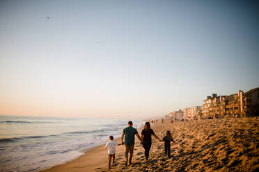 Vierköpfige Familie geht am Strand spazieren und hält sich bei Sonnenuntergang an den Händen - CAVF72751