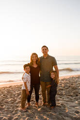 Vierköpfige Familie lächelt in die Kamera und umarmt sich am Strand bei Sonnenuntergang - CAVF72752