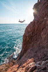 Young male jumps from red sand stone cliff into blue ocean - CAVF72889