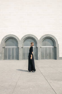 Junge Frau in langem schwarzen Kleid posiert vor den Türen des Gebäudes - CAVF72913