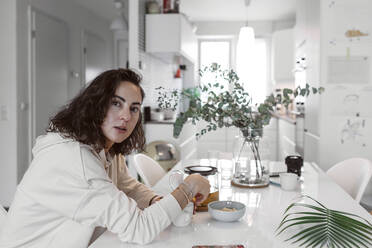 Portrait of woman sitting at kitchen table drinking tea - KMKF01196