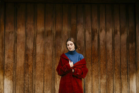 Nahaufnahme einer Frau mit blauem Rollkragenpullover und rotem Mantel vor einer Holztür, lizenzfreies Stockfoto