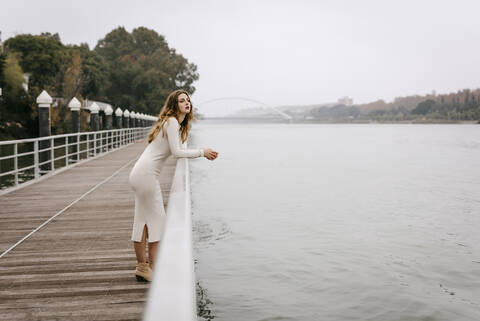 Porträt einer jungen Frau im weißen Kleid, die sich an einem regnerischen Tag an ein Geländer lehnt, lizenzfreies Stockfoto