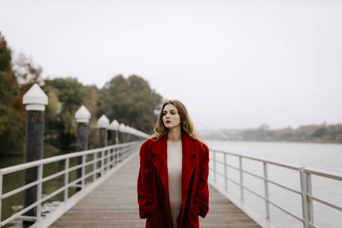 Porträt einer jungen Frau mit rotem Mantel auf einer Brücke an einem regnerischen Tag - TCEF00025
