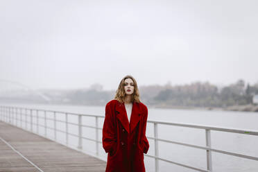 Portrait of young woman wearing red coat on a bridge during rainy day - TCEF00026