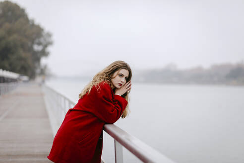 Porträt einer jungen Frau in rotem Mantel, die sich an einem regnerischen Tag an ein Geländer lehnt - TCEF00027