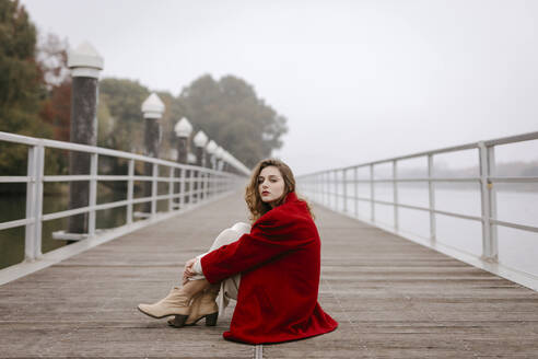 Porträt einer jungen Frau in rotem Mantel, die sich an einem regnerischen Tag an ein Geländer lehnt - TCEF00029
