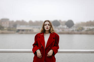 Portrait of young woman wearing red coat, leaning on railing during rainy day - TCEF00030