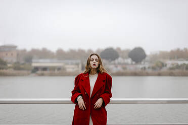 Portrait of young woman wearing red coat, leaning on railing during rainy day - TCEF00031