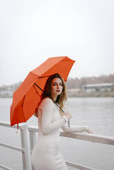 Porträt einer jungen Frau mit rotem Regenschirm, die sich an einem regnerischen Tag an ein Geländer lehnt - TCEF00032