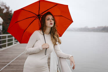 Porträt einer jungen Frau mit rotem Regenschirm, die sich an einem regnerischen Tag an ein Geländer lehnt - TCEF00034