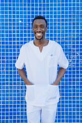 Porträt eines lachenden Arztes, der mit den Händen in den Taschen an einer blau gekachelten Wand steht - JCMF00347