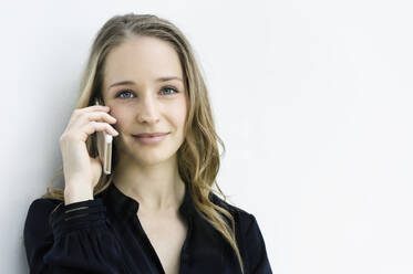 Junge Geschäftsfrau, die mit ihrem Smartphone vor einer weißen Wand spricht - CUF54459