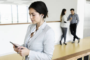 Junge Geschäftsfrau, die im Büro ihr Smartphone benutzt, im Hintergrund sprechen Kollegen - CUF54502