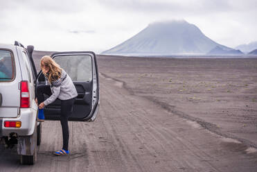 Woman changing shoes by vehicle door on dirt track, Landmannalaugar, Iceland - CUF54550