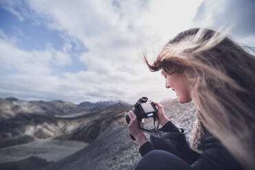 Female photographer with wind blown hair taking photograph on edge of volcano, Landmannalaugar, Iceland - CUF54562