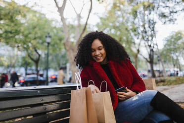 Lächelnde junge Frau schaut auf einer Parkbank in Einkaufstüten - FSIF04529