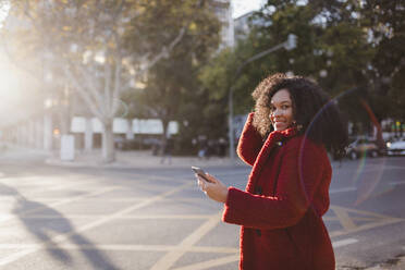 Porträt einer selbstbewussten, lächelnden jungen Frau mit Smartphone auf einer sonnigen Straße in der Stadt - FSIF04540