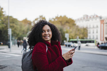 Smiling young woman with smart phone looking over shoulder on city street - FSIF04543